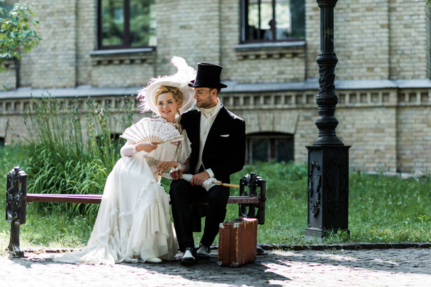cheerful woman holding fan while sitting with handsome victorian man on bench  - Fotografie, Obrázek