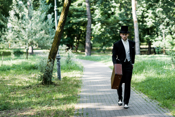 handsome victorian man in hat holding baggage and walking outside  - Фото, изображение