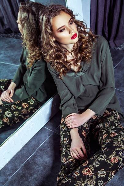 Mode-Modell mit Make-up und welliger Frisur lehnt sich an Spiegel und sitzt auf dem Boden in der Umkleidekabine  - Foto, Bild