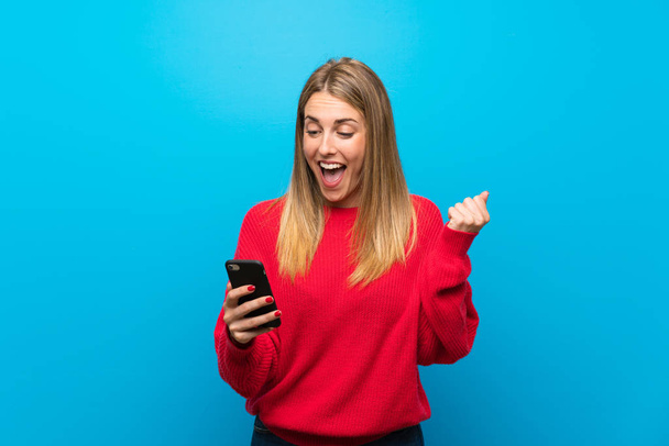 Mujer con suéter rojo sobre pared azul con teléfono en posición de victoria
 - Foto, Imagen
