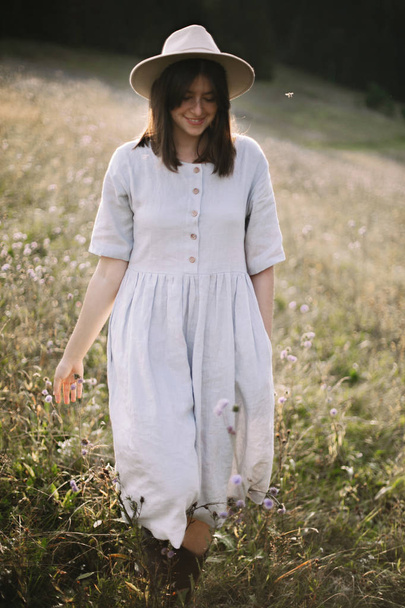 Stylish girl in rustic dress and hat walking among wildflowers a - Zdjęcie, obraz
