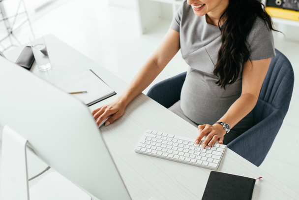 abgeschnittene Ansicht einer schwangeren Frau, die hinter einem Tisch sitzt und am Computer arbeitet - Foto, Bild