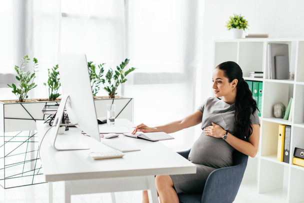 έγκυος γυναίκα κάθεται στο δωμάτιο με βιβλιοθήκη και γλάστρα με τα φυτά πίσω από το τραπέζι και εργάζονται σε υπολογιστή - Φωτογραφία, εικόνα