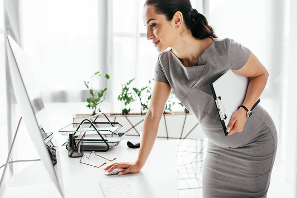 pregnant woman standing near table, holding clipboard and working on computer - Photo, Image