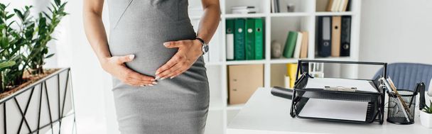 oříznutý pohled na těhotnou ženu přidržovací břicho při stání v kanceláři s květináčem a s knižní skříň poblíž stolu s podaváčem - Fotografie, Obrázek