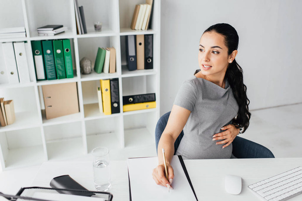 беременная женщина делает заметки, сидя за столом в офисе и отводя взгляд
 - Фото, изображение