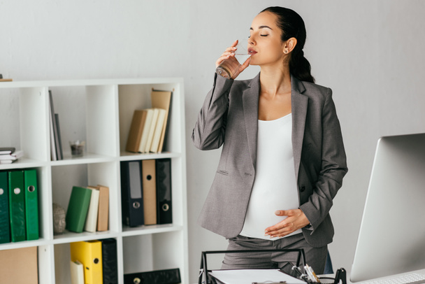pregnant woman standing near table and bookcase and drinking water from glass - Photo, Image