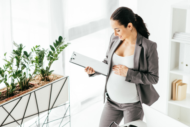 vista de ángulo alto de la mujer embarazada mirando a la carpeta mientras está de pie en la oficina cerca de maceta con planta
 - Foto, Imagen