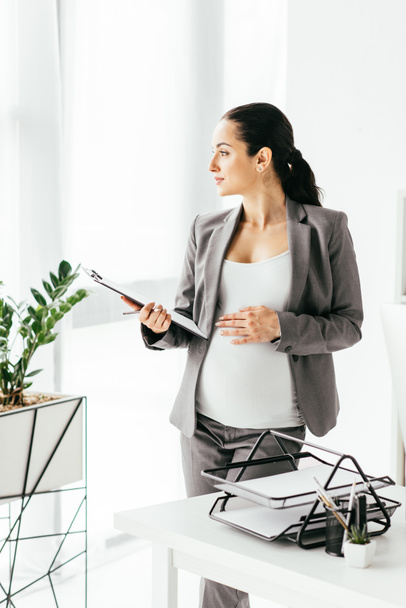 femme enceinte tenant ventre et dossier tandis que debout dans le bureau près de pot de fleurs et table avec plateau de documents et regarder loin
 - Photo, image