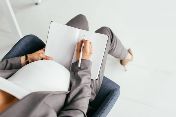 περικομμένη προβολή έγκυο γυναίκα Λαμβάνοντας σημειώσεις στο σημειωματάριο, ενώ κάθονται στο γραφείο σε καρέκλα γραφείου  - Φωτογραφία, εικόνα
