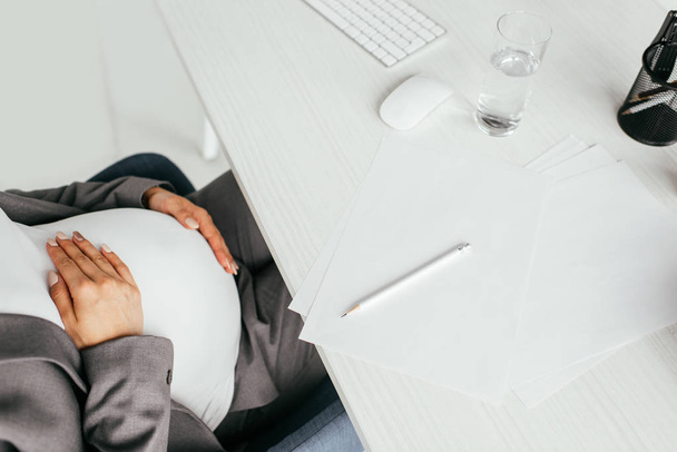oříznutý pohled na těhotnou ženu, která sedí za stolem se sklem, klávesnicí, papíry a tužkou - Fotografie, Obrázek