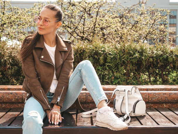 Portrait of beautiful brunette model dressed in summer hipster jacket and jeans clothes. Trendy girl sitting on the bench in the street background. Funny and positive woman in round sunglasses - Foto, Imagem