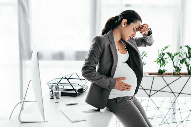 exhausted pregnant woman sitting on table, holding belly and enduring pain - Foto, afbeelding