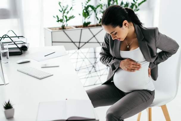 pregnant woman sitting in chair behind table in office, holding belly with hands and enduring pain - Photo, Image
