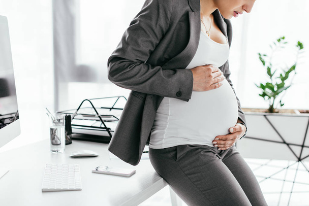 vue recadrée de femme enceinte tendue tenant et regardant le ventre tout en se tenant debout dans le bureau près de pot de fleurs
 - Photo, image