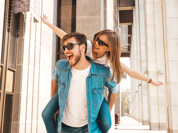 Smiling beautiful girl and her handsome boyfriend in casual summer clothes. Man carrying his girlfriend on the back and she raising her hands.Happy cheerful family having fun on the street background  - Foto, imagen