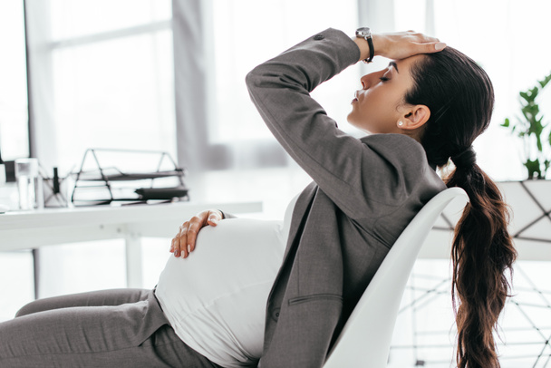 zmęczony Kobieta w ciąży siedzi w biurze w krzesło w pobliżu stołu i trzymając usłyszeć ręką - Zdjęcie, obraz