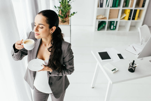 υψηλή γωνία θέα της εγκύου γυναίκα στέκεται στο γραφείο και πίνοντας καφέ  - Φωτογραφία, εικόνα