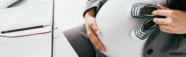 tiro panorâmico de mulher grávida segurando pequenas botas de bebê enquanto sentado atrás da mesa com notebook
 - Foto, Imagem