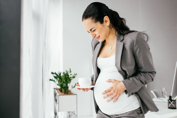 pregnant woman standing in office with cup of coffe and enduring pain - Fotoğraf, Görsel