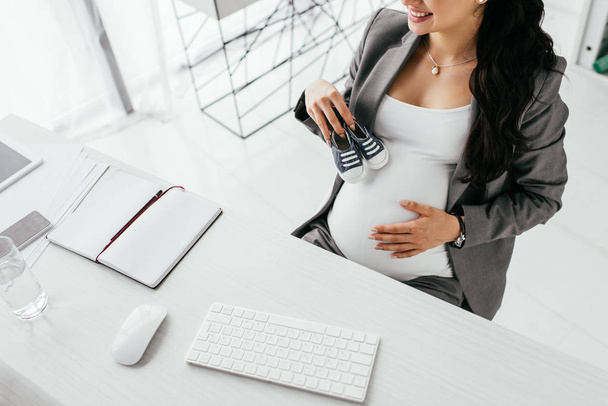 cropped view of pregnant woman sitting behind table with computer keyboard and mouse and holding small gumshoes - Foto, Imagen