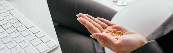 panoramic shot of woman holding colorful pills while sitting behind table - Photo, Image