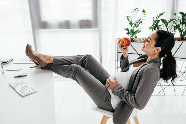 side view of pregnant woman holding apple while sitting on office - Foto, imagen
