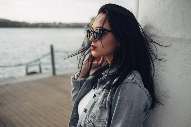 Beautiful Brunette Girl in Sunglasses White Pants and Jeans Jacket Near the Lake - Foto, afbeelding