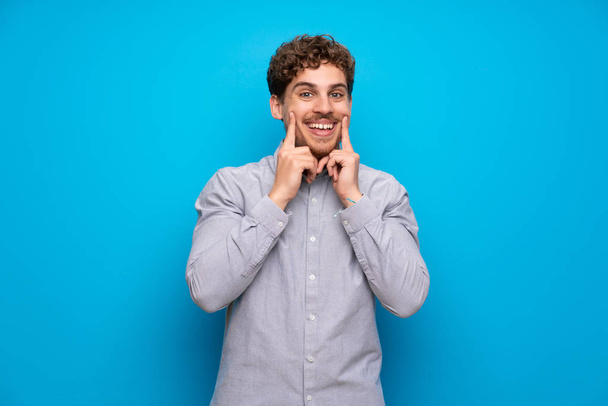 Blonde man over blue wall smiling with a happy and pleasant expression - Photo, Image