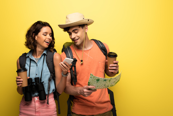 黄色の背景に地理マップを持つ男性にスマートフォンを示す陽気な女性 - 写真・画像