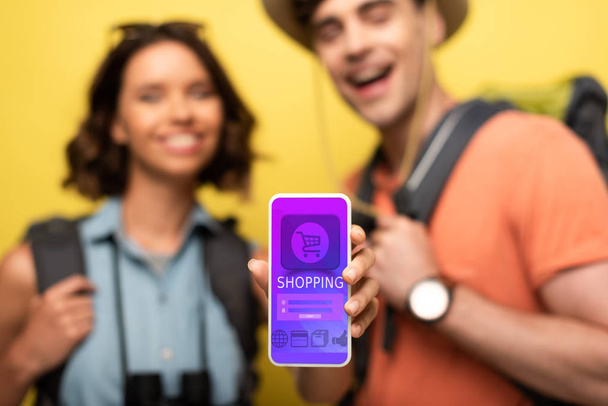 selective focus of cheerful woman showing smartphone with shopping app while standing near smiling man on yellow background - Foto, Bild