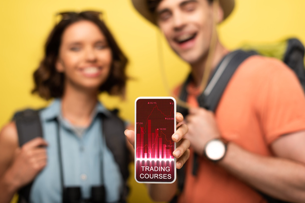 селективный фокус веселой женщины, показывающей смартфон с торговым приложением курсов, стоя рядом с улыбающимся мужчиной на желтом фоне
 - Фото, изображение