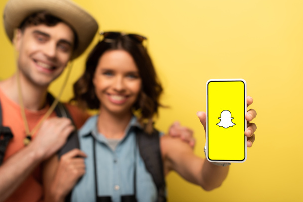 キエフ、ウクライナ - 2019年6月3日:陽気なボーイフレンドの近くに立って、黄色の背景にSnapchatアプリでスマートフォンを示す笑顔の女の子の選択的な焦点. - 写真・画像