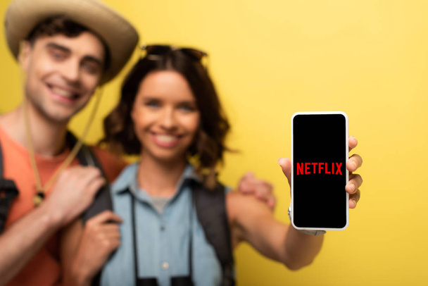 KYIV, UKRAINE - JUNE 3, 2019: Selective focus of cheerful young woman standing near smiling boyfriend and showing smartphone with Netflix app on yellow background. - Fotó, kép