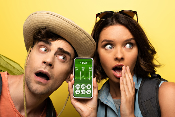 geschokt jonge vrouw tonen smartphone met Trading heartbeat rate app terwijl staande in de buurt van verrast man op gele achtergrond - Foto, afbeelding