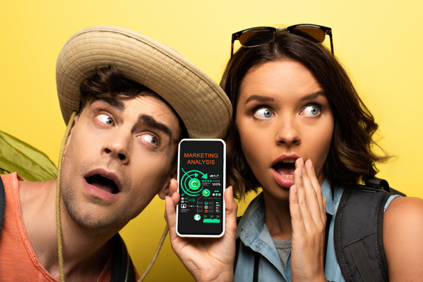 shocked young woman showing smartphone with marketing analysis app while standing near surprised man on yellow background - Photo, Image