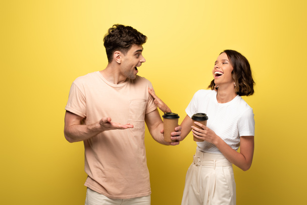excited man and woman talking while holding coffee to go on yellow background - 写真・画像