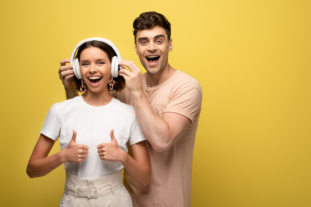 cheerful man putting on headphones on excited girl while looking at camera together on yellow background - Фото, зображення