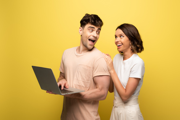 sarı arka planda neşeli kız yakınında dururken dizüstü bilgisayar kullanarak mutlu genç adam - Fotoğraf, Görsel