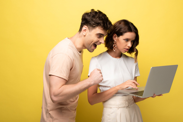 happy young man standing near attentive girl using laptop on yellow background - Fotoğraf, Görsel