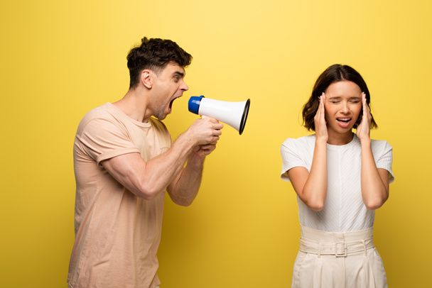angry young man quarreling in loudspeaker at girfriend covering ears with hands on yellow background - Fotoğraf, Görsel