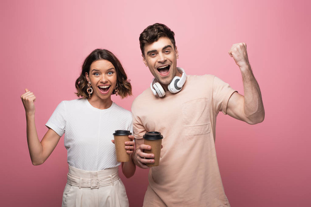 excited man and woman showing yes gestures while holding paper cups and looking at camera on pink background - Photo, image
