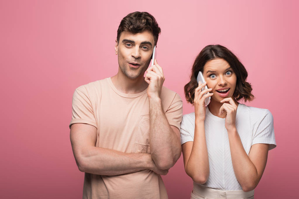 cheerful man and woman talking on smartphones while looking at camera on pink background - Photo, Image