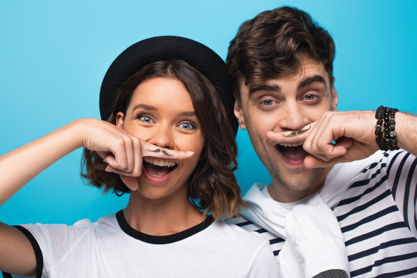 cheerful man and woman holding fingers with drawn mustache near faces on blue background - Fotoğraf, Görsel