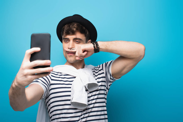 cheerful man taking selfie with smartphone while holding finger with drawn mustache near face on blue background - Foto, afbeelding