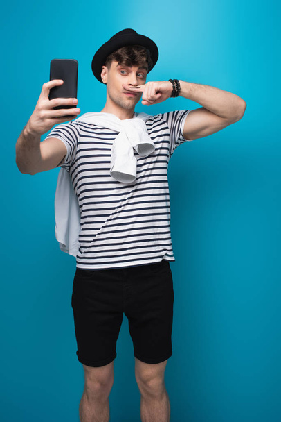 hombre guapo tomando selfie con teléfono inteligente mientras sostiene el dedo con bigote dibujado cerca de la cara sobre fondo azul
 - Foto, Imagen