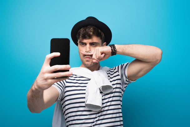 tricky man taking selfie with smartphone while holding finger with drawn mustache near face on blue background - Photo, Image