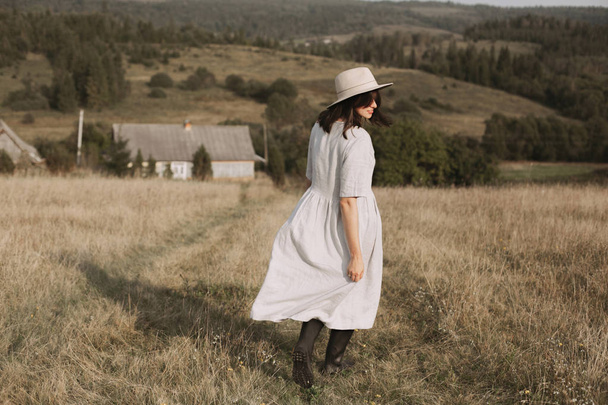 stilvolles Mädchen in Leinenkleid und Hut, das im sonnigen Feldgras spaziert - Foto, Bild