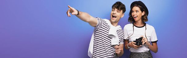 panoramic shot of cheerful man looking away and pointing with finger near smiling girl with digital camera on blue background - Photo, Image