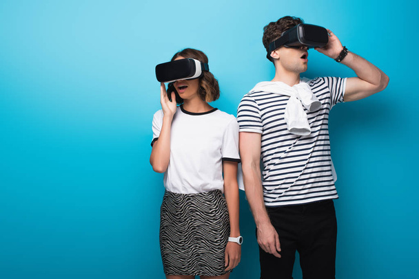 mladý, módní muž a žena, kteří používají sluchátka virtuální reality na modrém pozadí - Fotografie, Obrázek